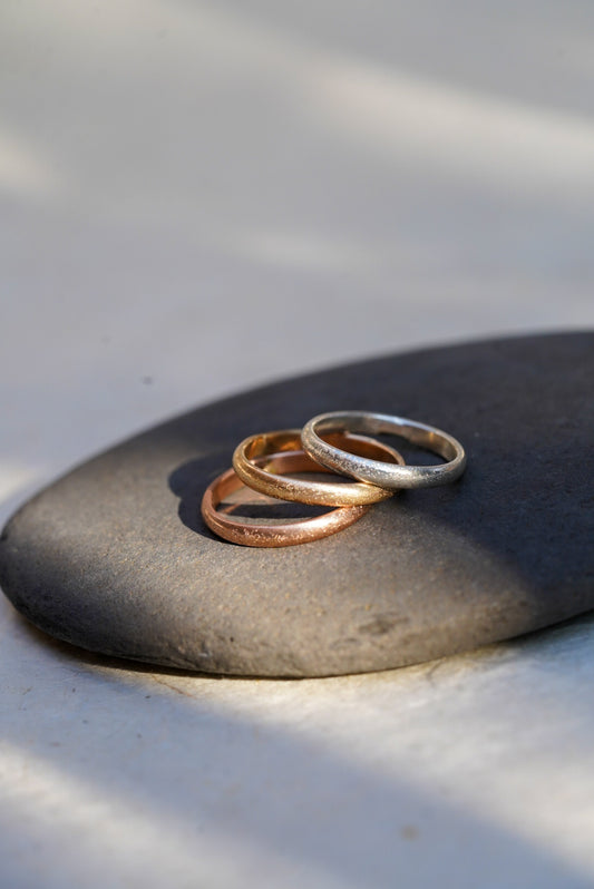 Three thin textured metal rings in silver, gold, and rose gold tones, stacked on a smooth dark gray stone against a light gray background with soft shadows.