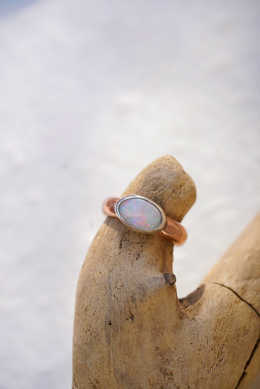 A rose gold ring with an oval cabochon opal gemstone, displayed on a piece of driftwood against a bright, snowy background.