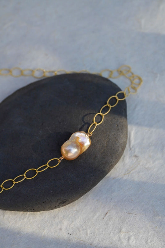 A delicate gold chain necklace featuring a single irregular, lustrous baroque pearl bead, displayed on a dark stone over a textured light gray surface.