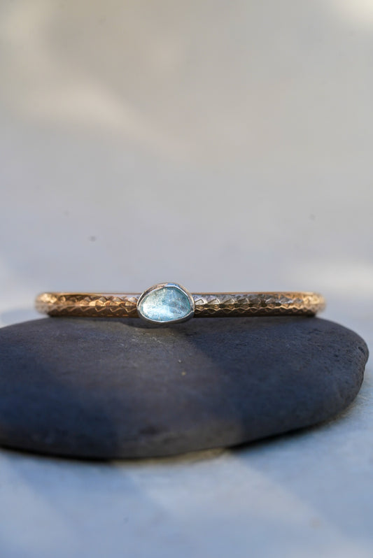 A delicate hammered gold bangle bracelet with a small, irregular-shaped light blue gemstone, resting on a smooth dark gray stone against a soft, light gray background.