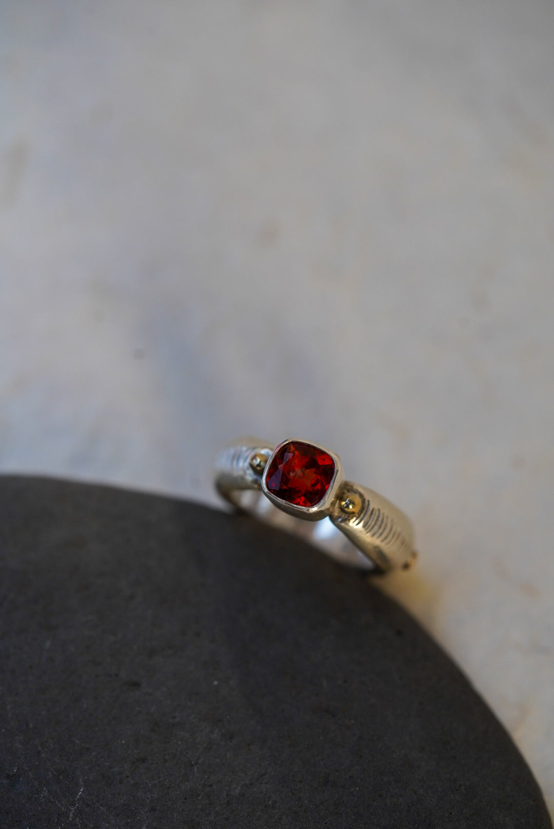 A silver ring with a square-cut red gemstone, displayed on a dark stone against a light marble background.