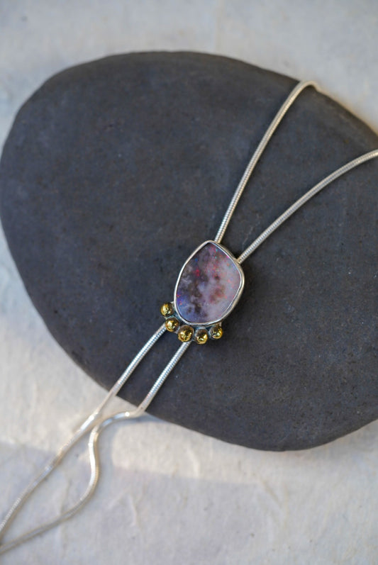 A close-up of a silver bolo necklace with a lavender and purple opal stone pendant, accented with small gold accents, resting on a dark gray stone against a light background.
