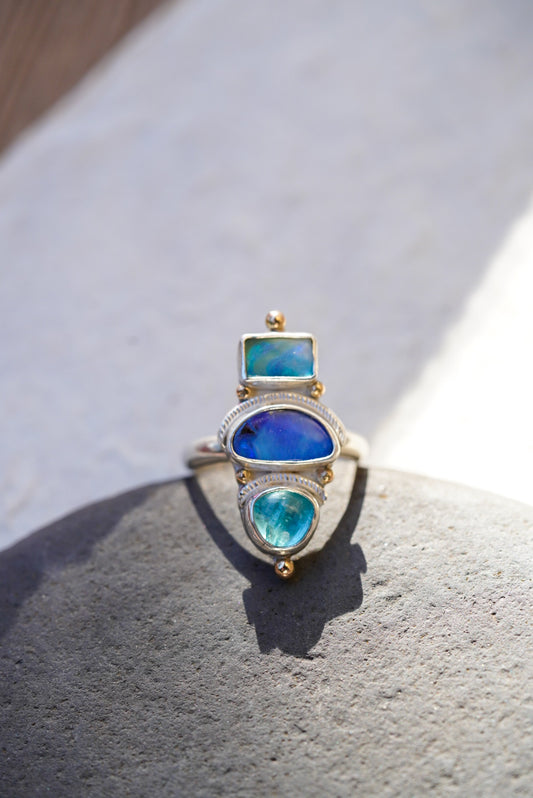 A silver ring with three stacked bright blue stones in vibrant blue and turquoise tones, displayed on a piece of rock against a neutral light background.