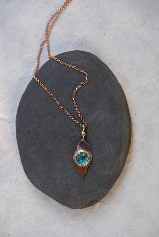 A rose gold necklace with a temple-shaped pendant featuring a round blue-green stone, displayed on a dark slate stone against a light textured background.