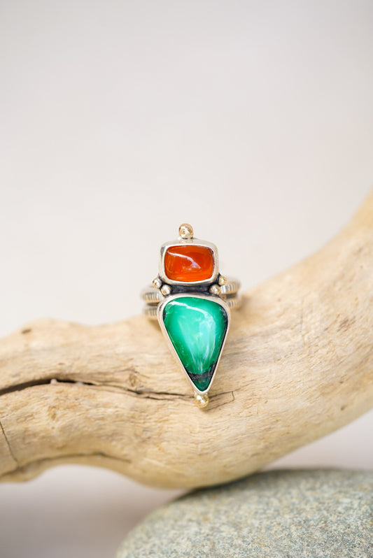 A silver ring featuring a large green teardrop-shaped variscite stone and a smaller orange oval chalcedony, displayed on a piece of driftwood against a light gray background.