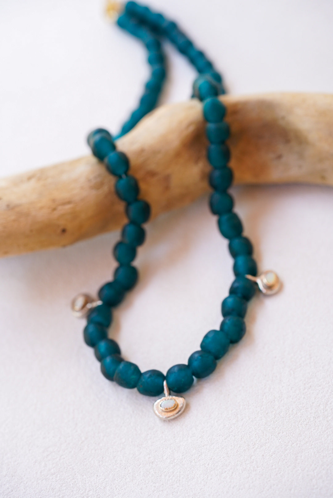 A beaded necklace made of dark teal-green beads with small silver oval charms with opals, draped over a piece of driftwood on a light textured background