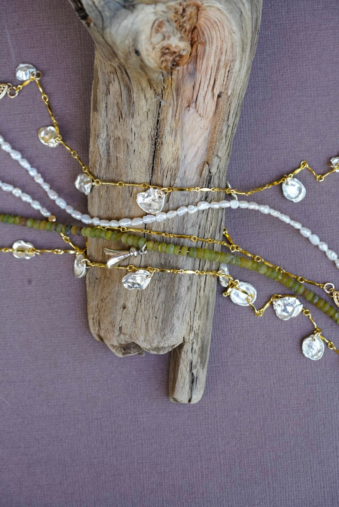 A close-up of delicate anklets draped over a piece of driftwood, featuring gold chains with recycled silver beads, white pearl strands, and olive green beads, set against a textured mauve background.