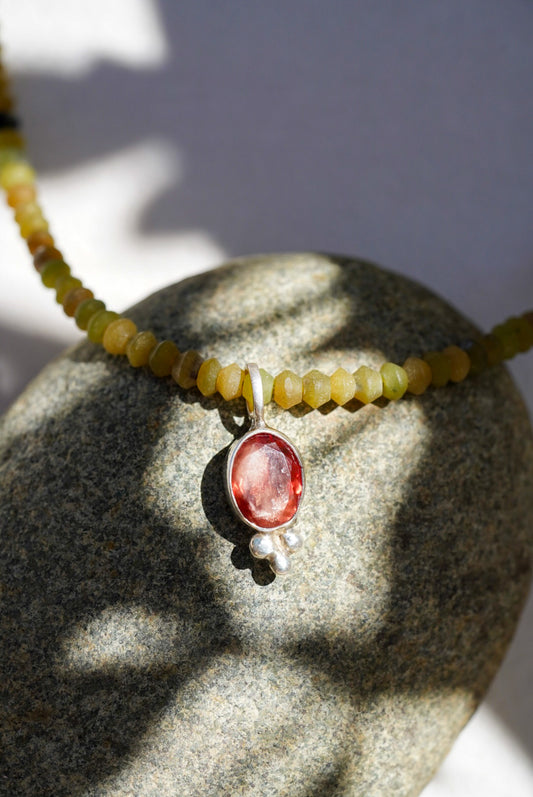 A close-up of a beaded necklace draped over a smooth gray stone, featuring small yellow-green beads and a central oval pendant with a pinkish-red stone