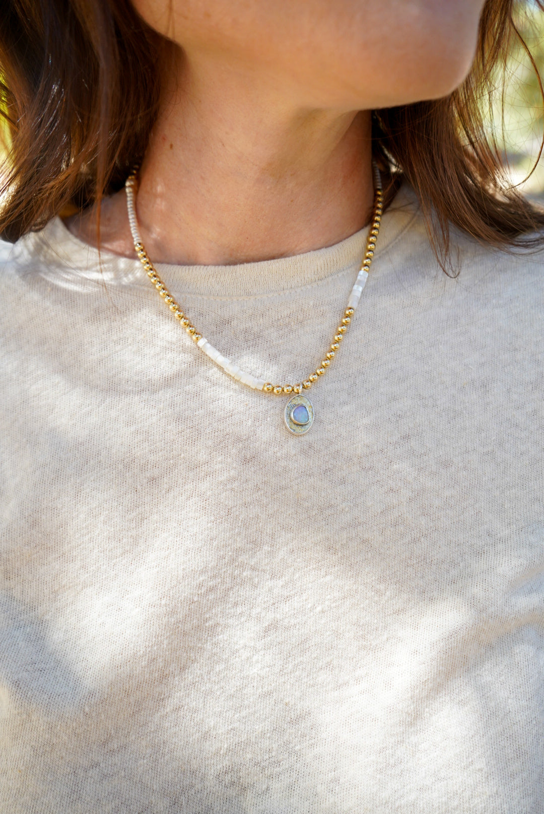 A close-up lifestyle shot of a person wearing a delicate gold beaded necklace with a small oval opal stone pendant, paired with a white pearl and gold beaded necklace.