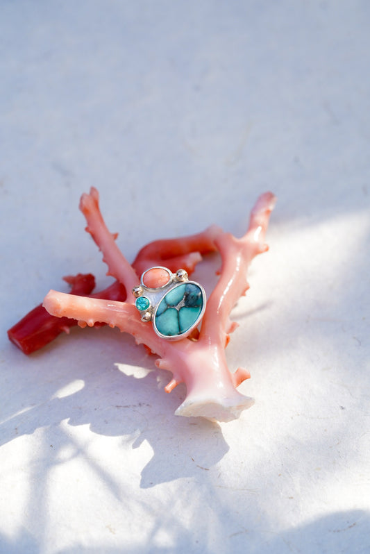 A silver ring with a large turquoise-blue oval stone and smaller coral accents, displayed on a pink coral fragment against a bright white background.