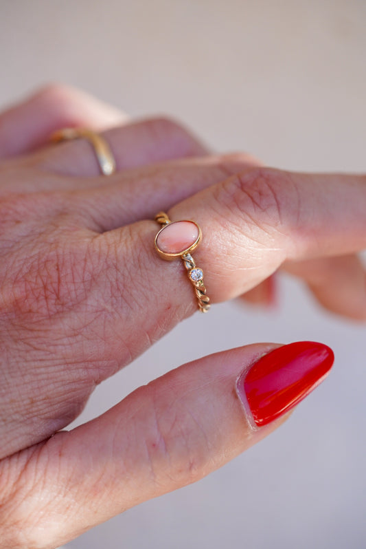 A close-up of a hand wearing a delicate gold ring with a small oval pink coral stone and diamond accent stone, alongside other rings and a red-painted fingernail.