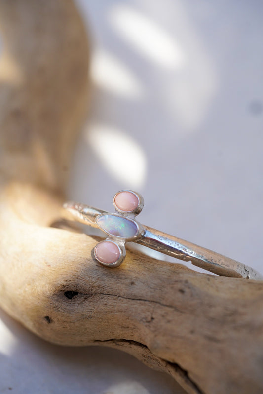 A silver cuff with a central iridescent opal and two small pink coral stones, displayed on a piece of driftwood.