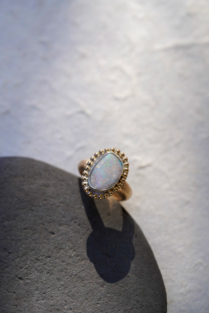 A gold ring with an iridescent white opal center stone, displayed on a dark textured stone against a light textured background.