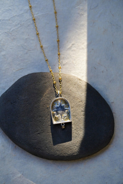 A gold chain necklace with a arched pendant containing a silver star with a dusting of gold, resting on a dark stone against a textured light background.
