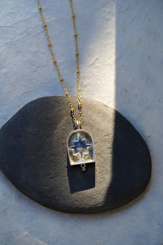 A gold chain necklace with a arched pendant containing a silver star with a dusting of gold, resting on a dark stone against a textured light background.