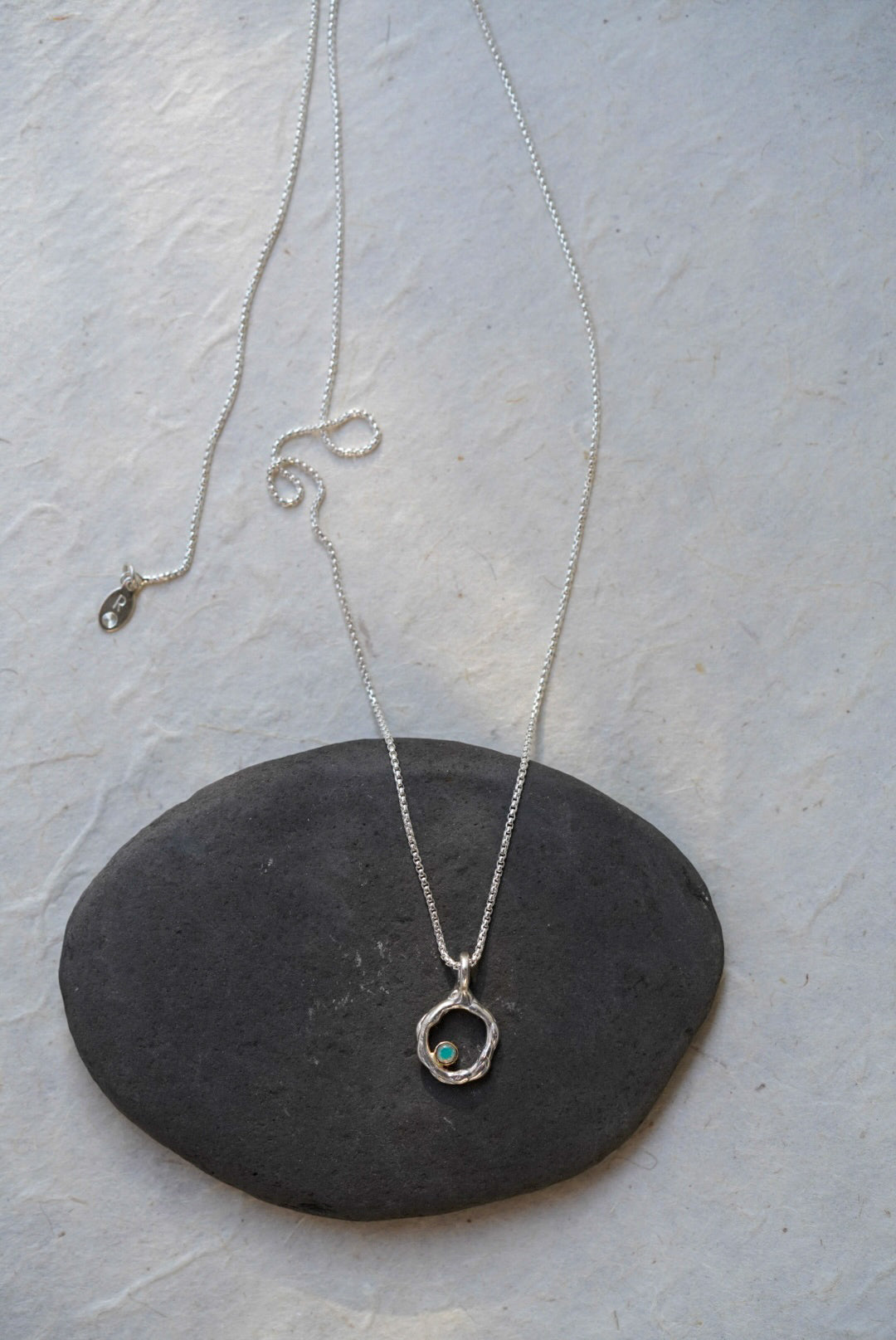A silver chain necklace with a circular openwork pendant and a small opal stone, displayed on a dark oval stone against a light textured background.