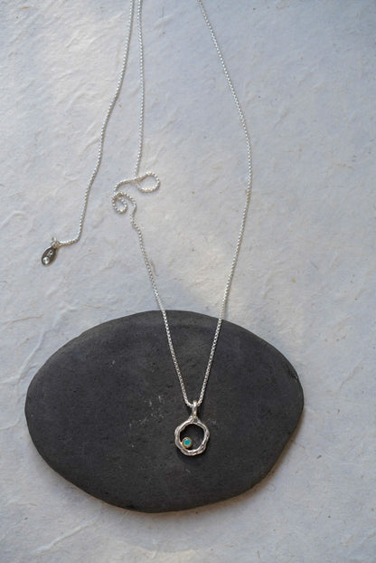 A silver chain necklace with a circular openwork pendant and a small opal stone, displayed on a dark oval stone against a light textured background.