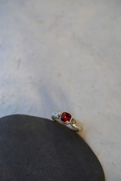 A silver ring with a cushion-cut red gemstone, displayed on a dark gray stone against a light marble background.