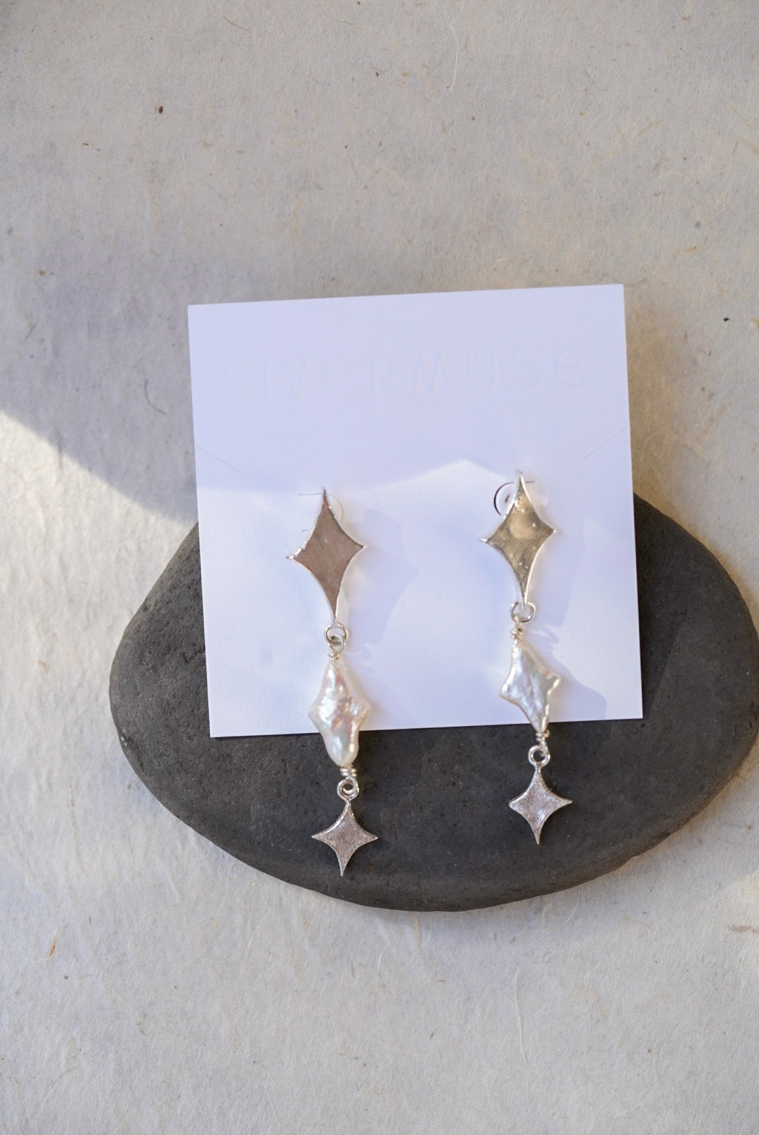A pair of silver dangle earrings with star-shaped and star pearls, displayed on a white card resting on a dark stone, photographed against a light textured wall.
