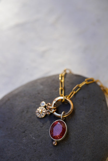 A gold chain necklace with a ring clasp and three dangling charms, including a faceted oval red sunstone, a sun and small Damond displayed on a dark gray stone against a light gray background.