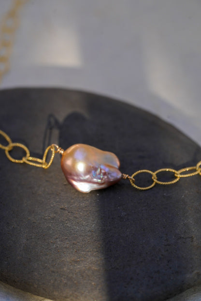 A close-up of a delicate gold chain necklace featuring a single baroque pearl pendant with iridescent pink, purple, and blue hues, resting on a dark textured stone surface.