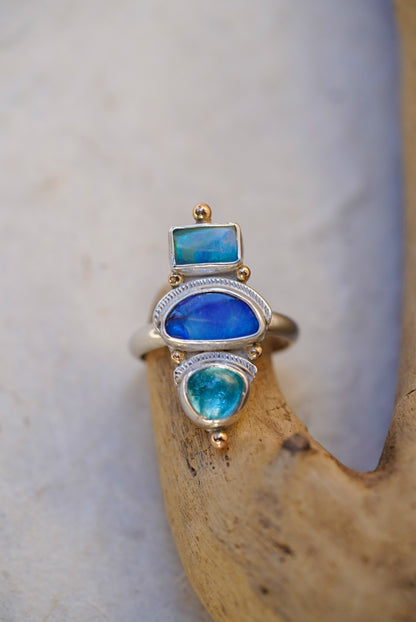 A silver ring with three stacked bright blue stones in vibrant blue and turquoise tones, displayed on a piece of driftwood against a neutral light background.