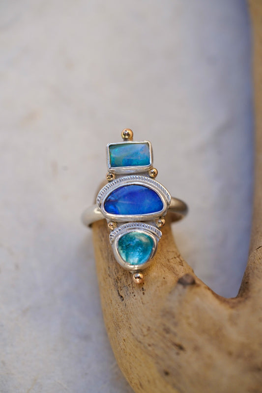 A silver ring with three stacked bright blue stones in vibrant blue and turquoise tones, displayed on a piece of driftwood against a neutral light background.