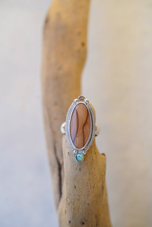 A silver ring with an oval cabochon stone and a small turquoise accent, displayed on a piece of driftwood against a neutral light background.