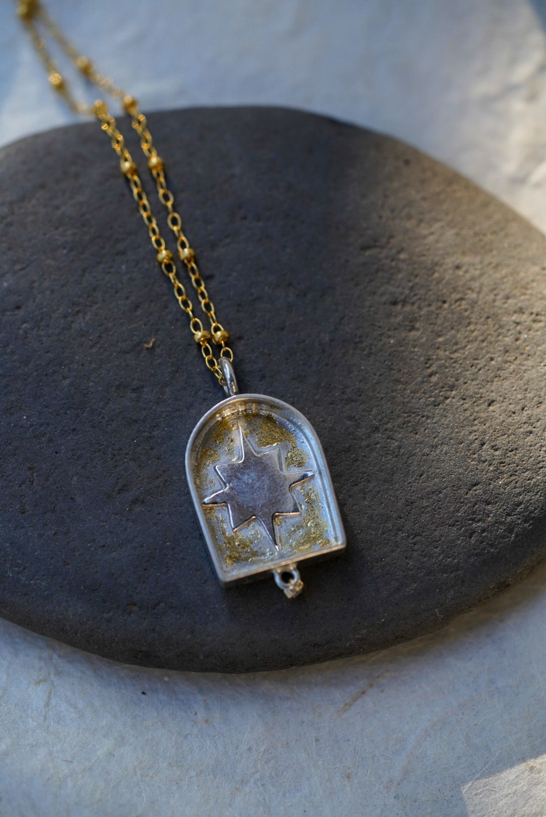 A gold chain necklace with a arched pendant containing a silver star with a dusting of gold, resting on a dark stone against a textured light background.