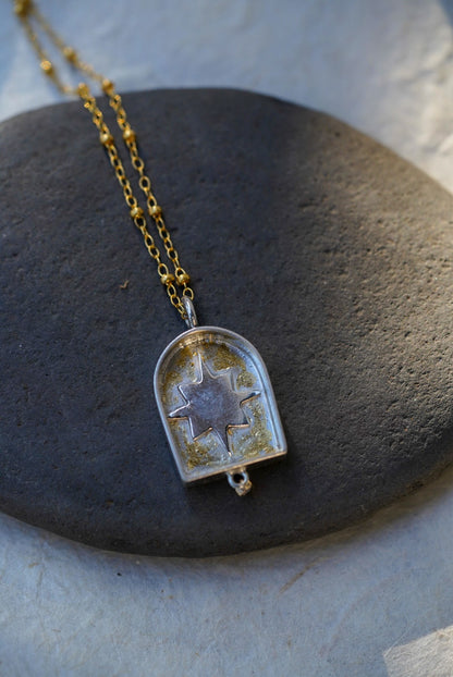 A gold chain necklace with a arched pendant containing a silver star with a dusting of gold, resting on a dark stone against a textured light background.