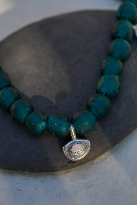 A close-up of a beaded necklace with dark teal-green round beads and a small silver-toned pendant featuring a pink and white opal, resting on a dark stone surface.