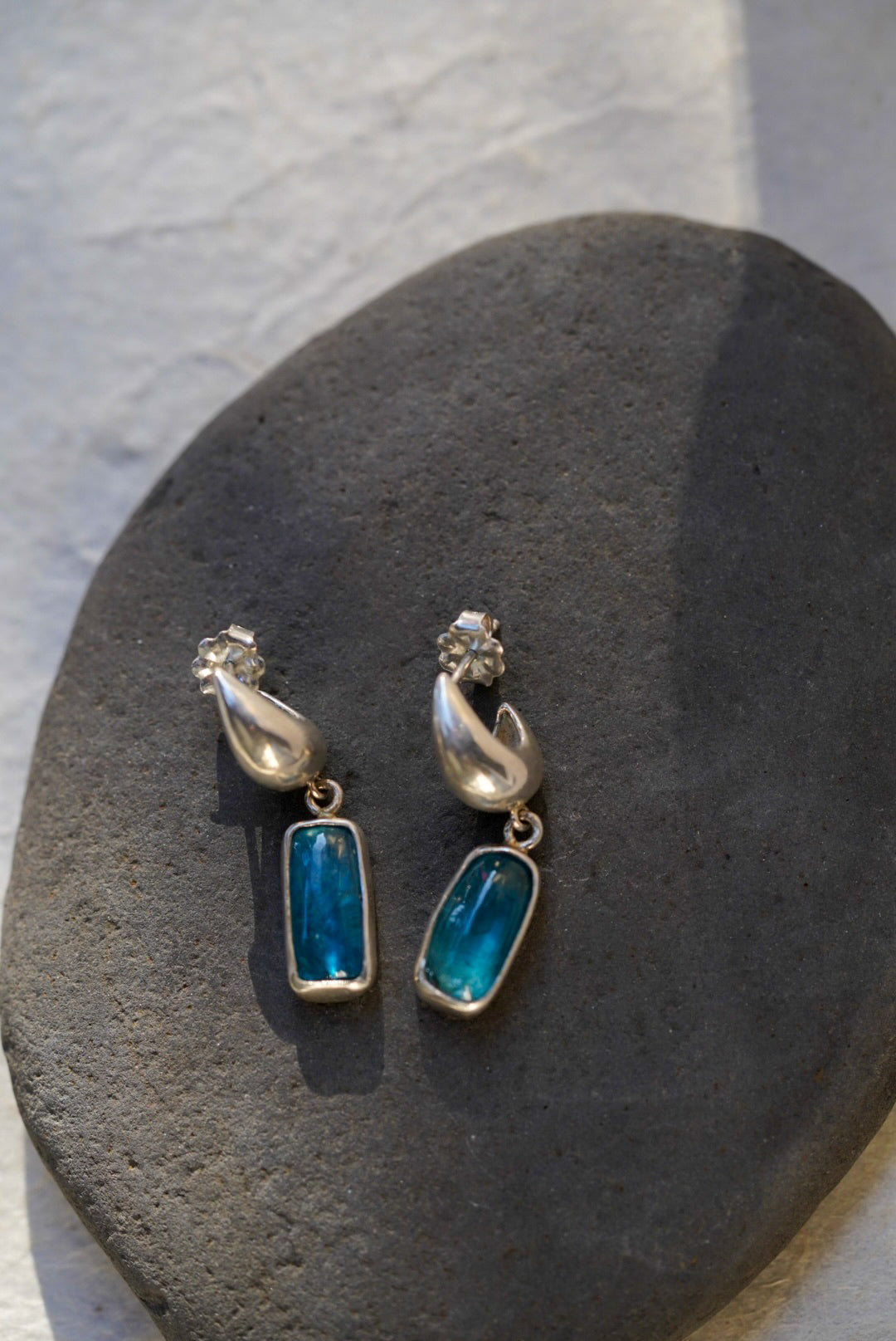 A pair of silver dangle earrings with rectangular blue-green stones, displayed on a dark gray stone against a light gray textured background.