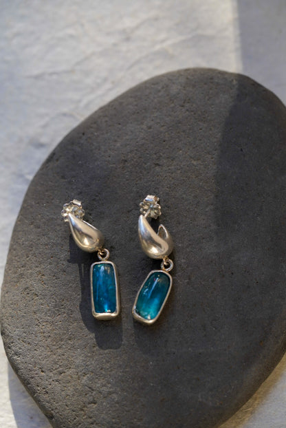 A pair of silver dangle earrings with rectangular blue-green stones, displayed on a dark gray stone against a light gray textured background.