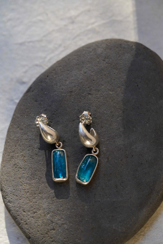 A pair of silver dangle earrings with rectangular blue-green stones, displayed on a dark gray stone against a light gray textured background.