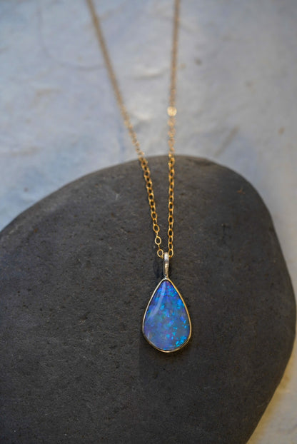 A gold chain necklace with a teardrop-shaped blue opal pendant, displayed on a dark gray stone against a light textured background.