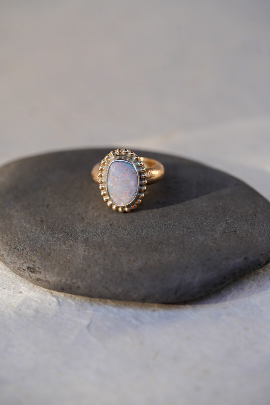 A gold ring with an iridescent white opal center stone, displayed on a dark textured stone against a light textured background.