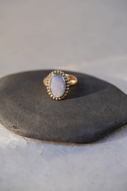 A gold ring with an iridescent white opal center stone, displayed on a dark textured stone against a light textured background.