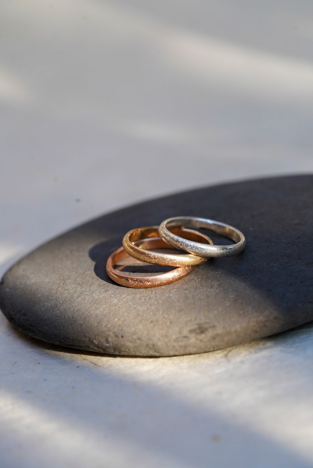 Three thin textured metal rings in silver, gold, and rose gold tones, stacked on a smooth dark gray stone against a light gray background with soft shadows.