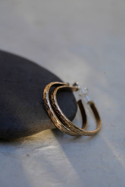 A pair of textured gold hoop earrings resting on a smooth dark stone against a light gray background.