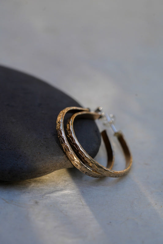 A pair of textured gold hoop earrings resting on a smooth dark stone against a light gray background.