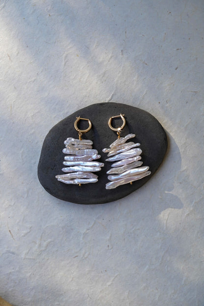 A pair of handmade dangle earrings with gold hoops and stacked iridescent white pearls, displayed on a dark stone against a textured light gray background.