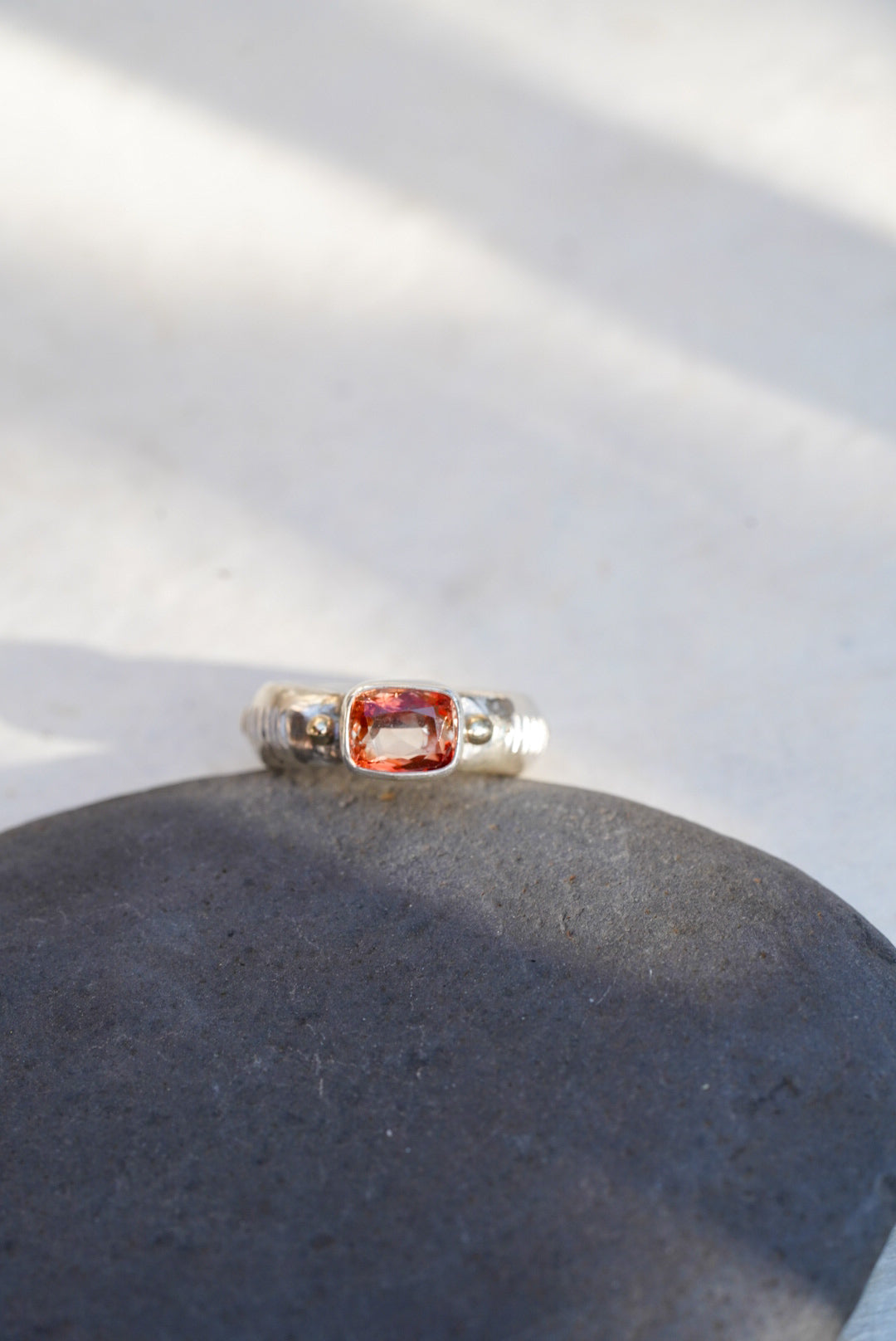 A silver ring with a rectangular orange-red gemstone, resting on a dark gray stone against a light background with soft shadows.