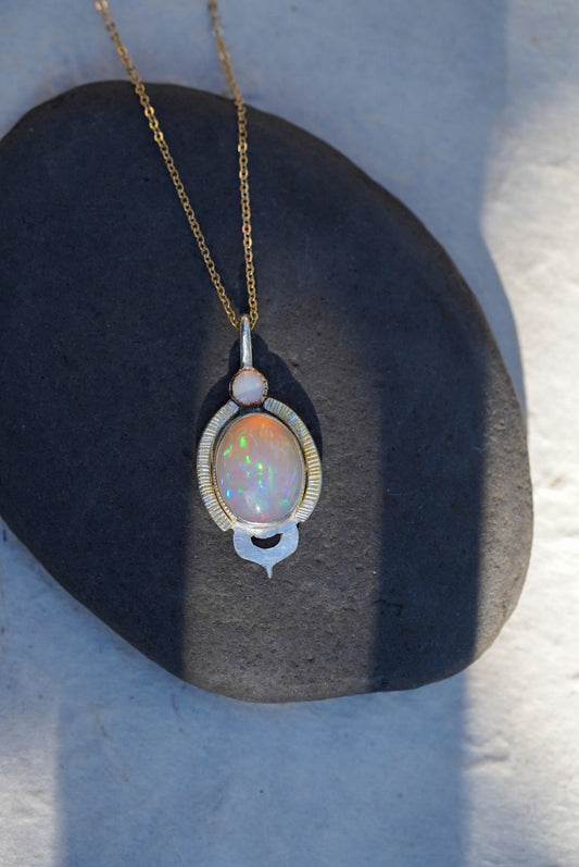 A gold chain necklace with an oval opal pendant, displayed on a dark stone against a light background.