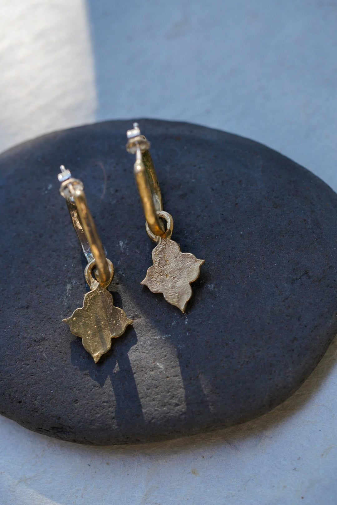 A pair of textured bronze temple charm drop earrings resting on a dark, smooth stone against a light gray background.