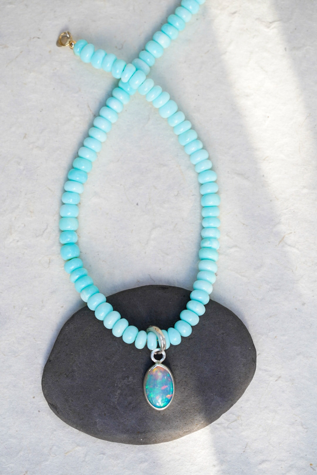 A baby blue beaded necklace with removable charm in rainbow opal, set upon a black rock on a light background.