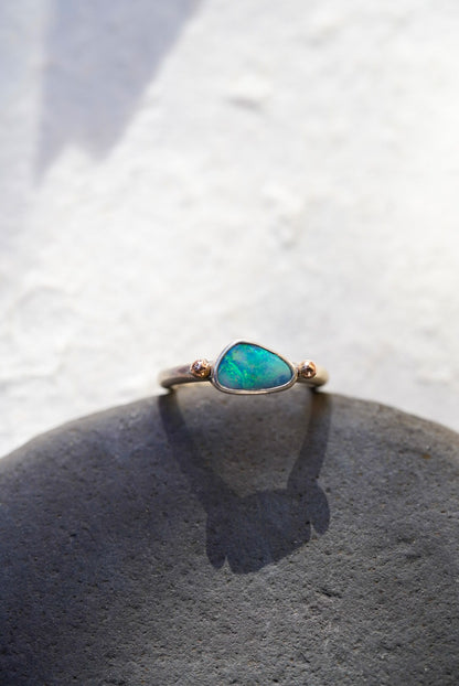 A delicate ring with a green-blue opal center stone and rose gold side accents, resting on a dark gray stone against a textured white background.
