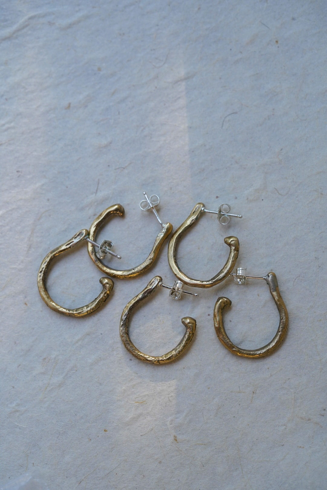 Four textured bronze hoop earrings with silver posts, arranged on a light beige textured paper background.