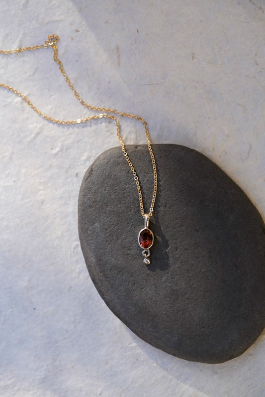 A delicate gold chain necklace with an oval amber-brown gemstone pendant and a small diamond accent stone, displayed on a dark gray stone slab against a light beige background.