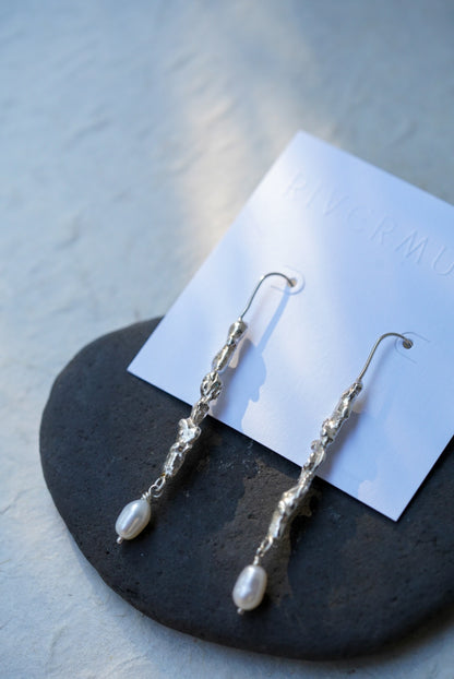 A pair of silver dangle earrings with white pearl drops, displayed on a white card over a dark stone surface.