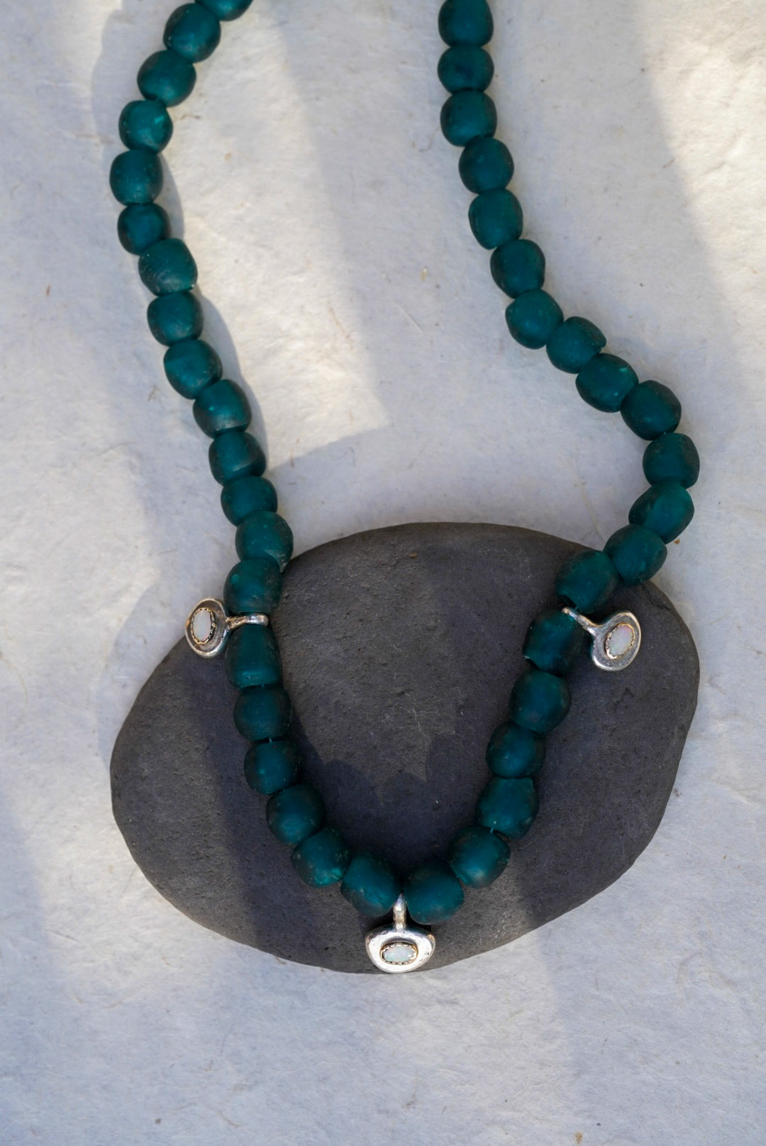 A beaded necklace made of dark teal-green beads with small silver oval charms with opals,  draped over a dark gray stone on a light textured background.